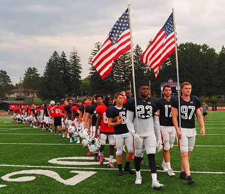 The football team walking on the field, carrying two American flags